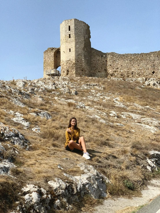 a woman sitting on a hill with a castle in the background, 🤠 using a 🖥, byzantine ruins, low quality photo, photo on iphone