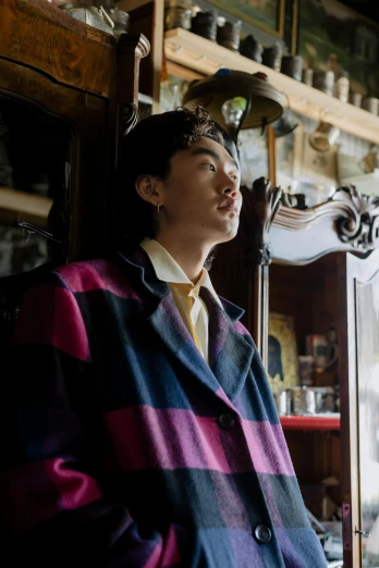a man standing next to a window in a room, an album cover, inspired by Zhang Han, unsplash, wearing a purple detailed coat, tartan garment, official store photo, androgynous person