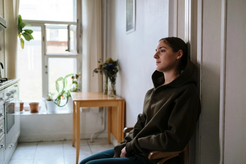 a woman sitting on a chair in a kitchen, pexels contest winner, realism, girl wearing hoodie, sad man, window light, sitting in a waiting room