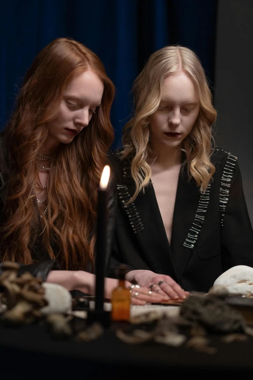 two women sitting at a table with a candle, an album cover, trending on pexels, renaissance, sophie turner, occult robes, ( ( theatrical ) ), skulls in hands