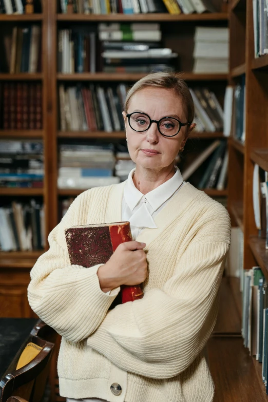 a woman standing in front of a bookshelf holding a book, by Grytė Pintukaitė, square rimmed glasses, high-quality photo, russian academic, proud serious expression