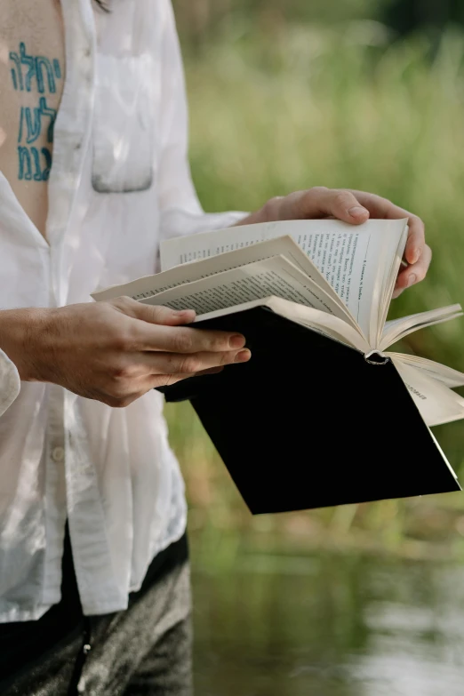 a woman holding a book near a body of water, in a field, photo of a man, black, opening