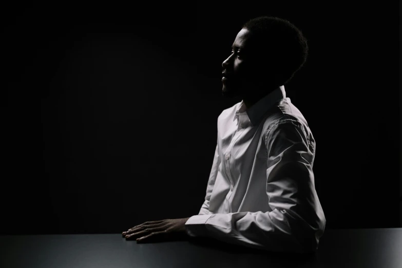 a man in a white shirt sitting at a table, by Clifford Ross, pexels contest winner, dark-skinned, elegant profile pose, screensaver, shot at dark with studio lights