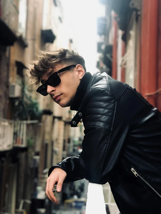 a man in a leather jacket leaning on a railing, an album cover, inspired by Luca Zontini, trending on pexels, wearing oakley sunglasses, profile image, androgynous face, in an alley