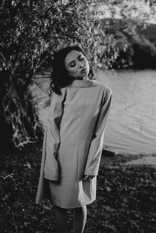 a woman standing next to a body of water, a black and white photo, inspired by irakli nadar, unsplash, ao dai, wearing a trenchcoat, wearing a nightgown, wearing a turtleneck and jacket