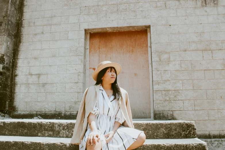 a woman sitting on the steps of a building, pexels contest winner, beige fedora, mai anh tran, simple cream dress, striped