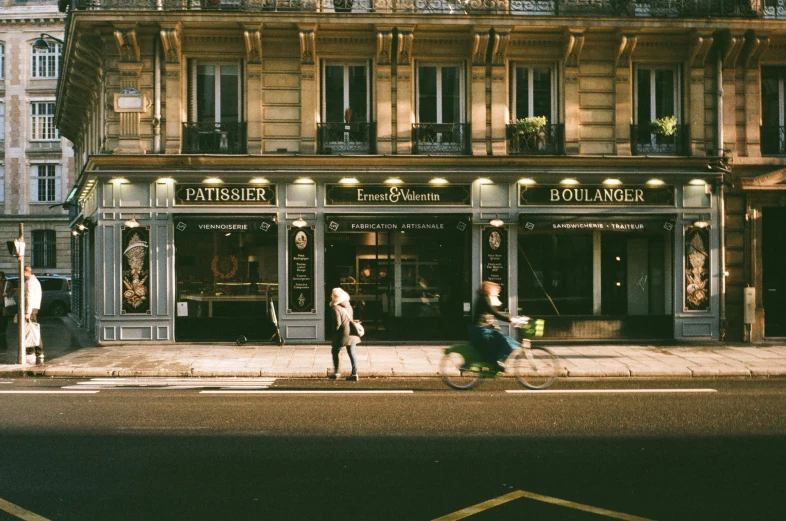 a man riding a bike down a street next to a tall building, pexels contest winner, neoclassicism, inside a french cafe, pur champagne damery, warm street lights store front, people walking around