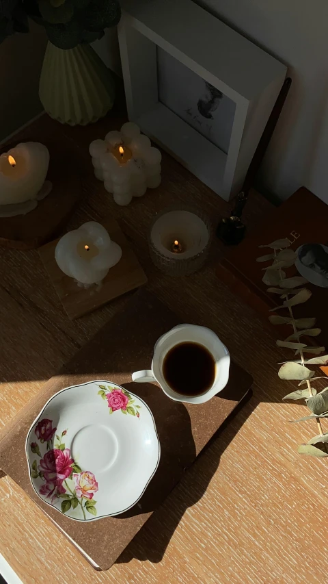 a cup of coffee sitting on top of a wooden table, a still life, inspired by Elsa Bleda, trending on pexels, room full of candles, soft shadow, a high angle shot, soft bloom lighting