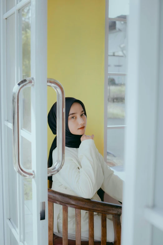 a woman sitting in a chair looking out a window, by Basuki Abdullah, trending on pexels, realism, white hijab, yellow and black, door, handsome girl