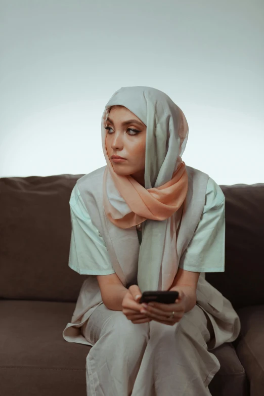 a woman sitting on a couch holding a cell phone, a colorized photo, inspired by Maryam Hashemi, trending on pexels, hurufiyya, concerned expression, white hijab, people watching, gen z