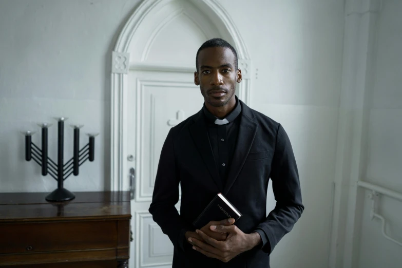 a man standing in a room holding a book, scene set in a church, dark-skinned, portrait image, dressed in black