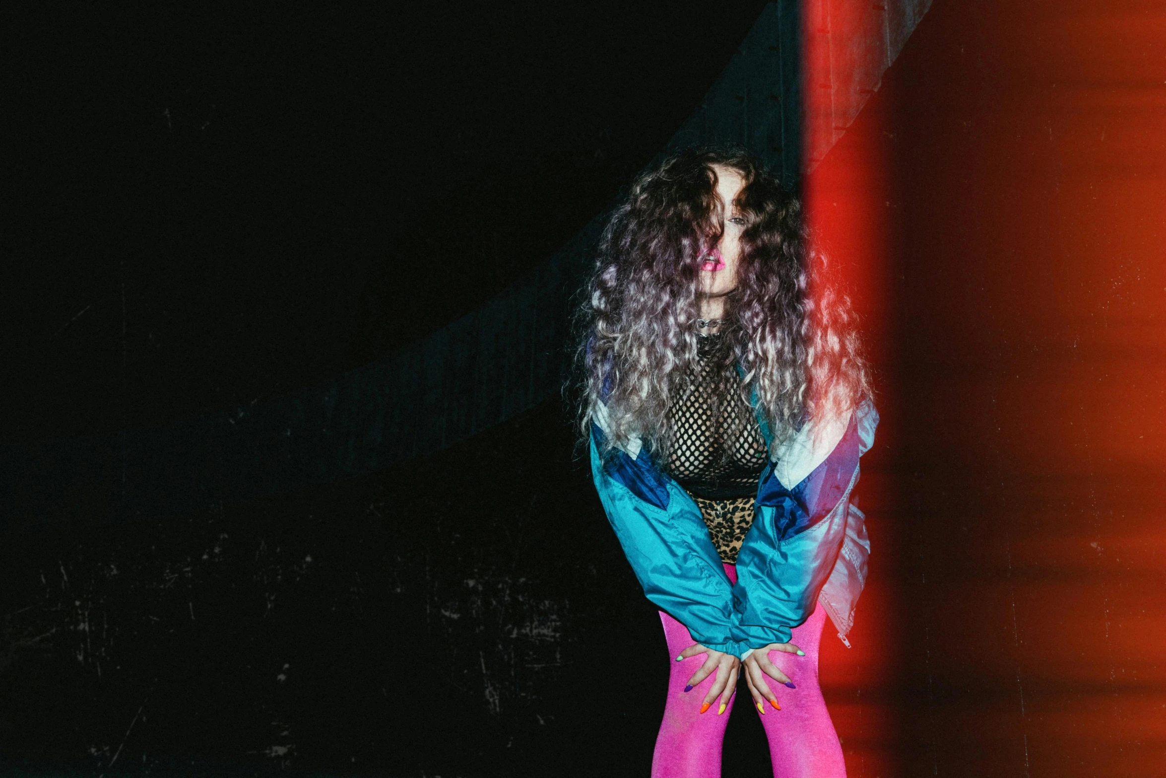 a woman standing in front of a red wall, an album cover, inspired by Elsa Bleda, pexels contest winner, graffiti, huge curly pink hair, crawling out of a dark room, holographic suit, purple and blue neon