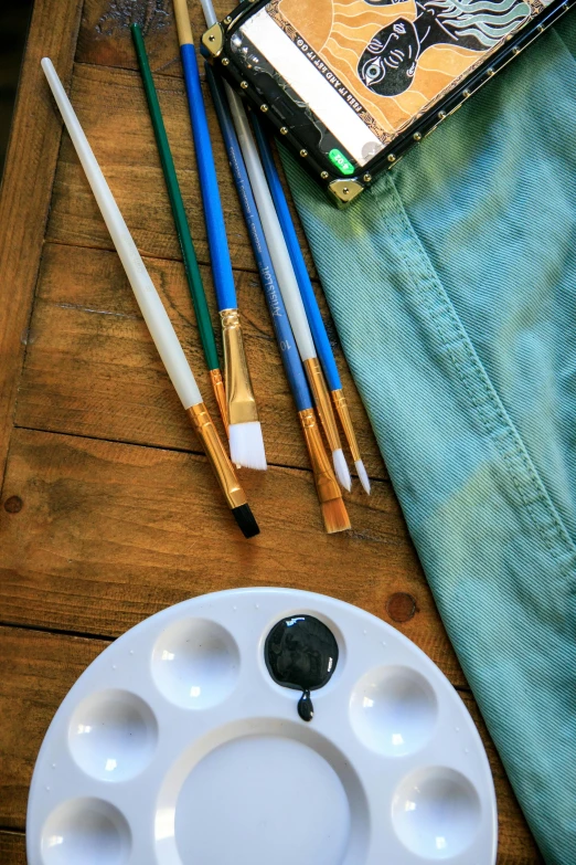 a wooden table topped with paint brushes and a palette, painted overalls, 🎨🖌, tie-dye, brown