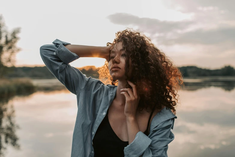 a woman standing next to a body of water, trending on pexels, happening, brown curly hair, magic hour lighting, redahair and attractive features, arm around her neck