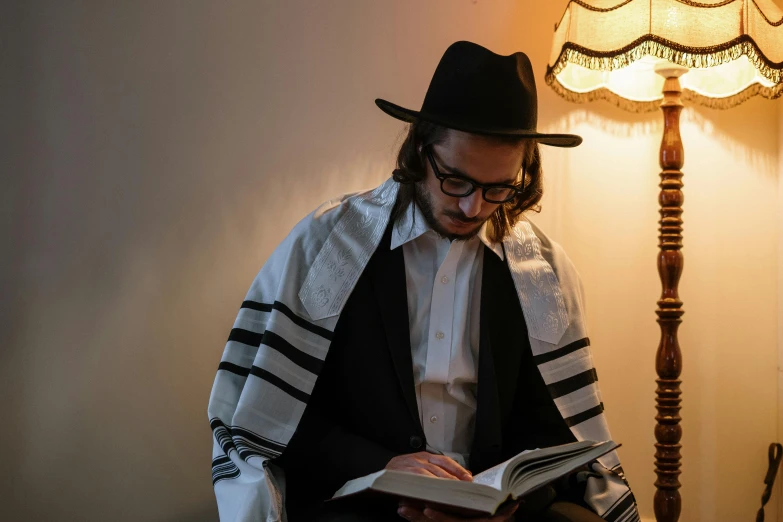 a man sitting in a chair reading a book, by Daniel Lieske, pexels, sukkot, he is casting a lighting spell, baggy clothing and hat, jewish young man with glasses