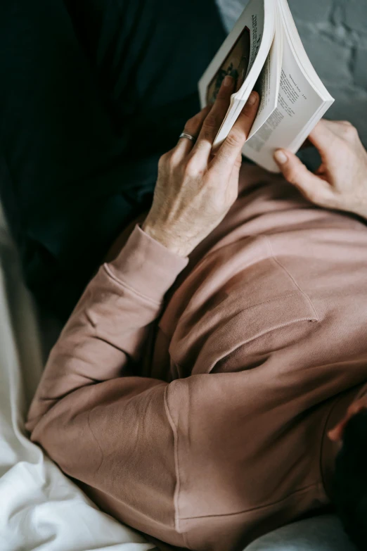 a man laying on a bed reading a book, by Grytė Pintukaitė, trending on unsplash, wearing a pastel pink hoodie, older woman, holding a leather purse, laying down with wrists together