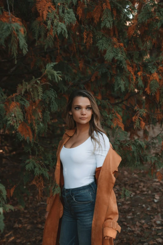 a woman standing in front of a tree, by Robbie Trevino, trending on pexels, brown jacket, isabela moner, she has long orange brown hair, an all white human