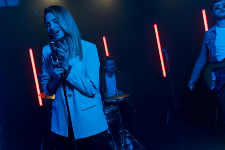 a woman standing next to a man holding a microphone, an album cover, pexels, antipodeans, neon operator margot robbie, band playing, blue light, thumbnail