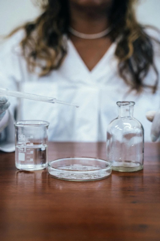 a woman in a lab coat sitting at a table, pexels, process art, glassware, environmental shot, college, white