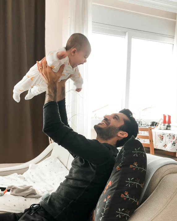 a man holding a baby up in the air, pexels contest winner, happening, sitting on the couch, bowater charlie and brom gerald, real life photo of a syrian man, cute and lovely