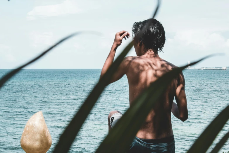 a man standing on top of a beach next to the ocean, pexels contest winner, visual art, chest covered with palm leaves, profile image, thawan duchanee, showing her shoulder from back