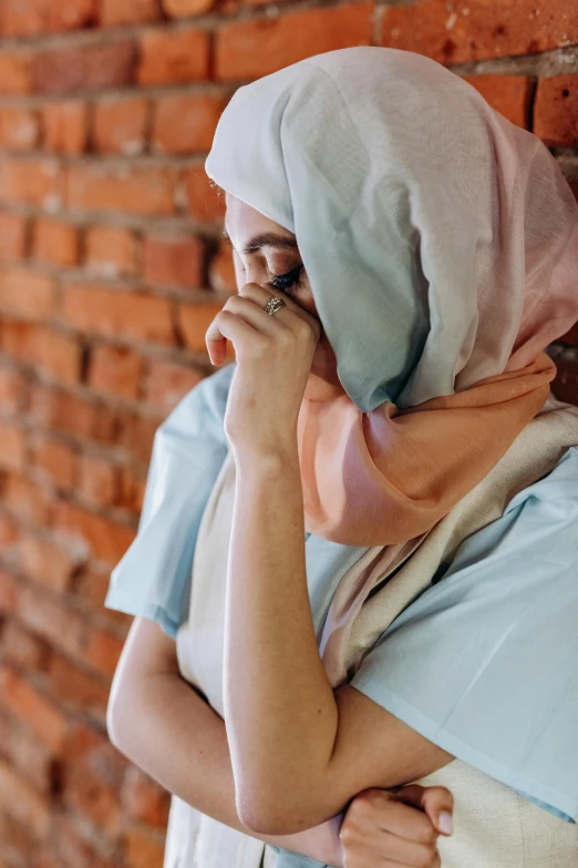 a woman standing in front of a brick wall talking on a cell phone, by Maryam Hashemi, pexels, symbolism, woman crying, wearing a long flowy fabric, pastel blues and pinks, praying posture