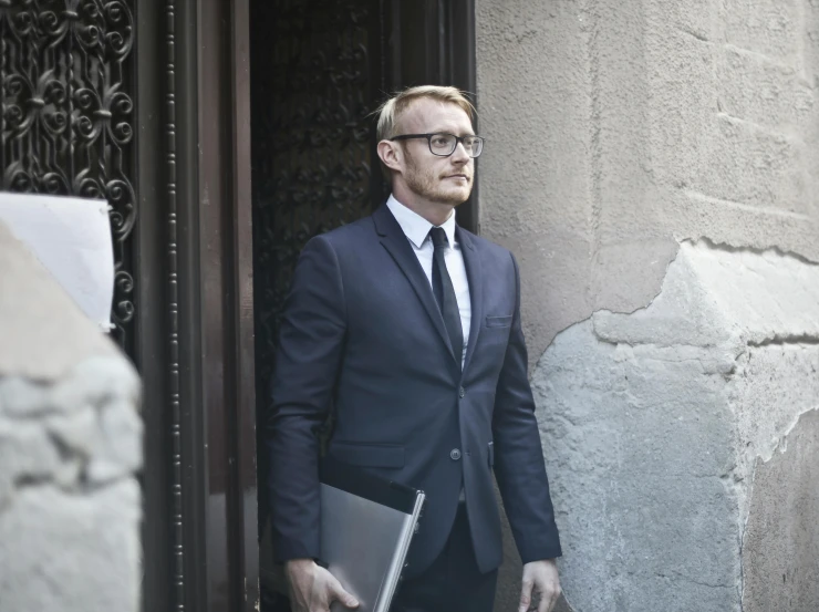 a man in a suit and tie holding a laptop, by Giuseppe Avanzi, pexels contest winner, renaissance, leaning on door, wearing black rimmed glasses, promotional image, hunting