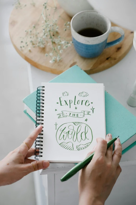 a person writing on a notebook next to a cup of coffee, inspired by Ruth Jên, sea green color theme, explorer sketch, thumbnail, calligraphy