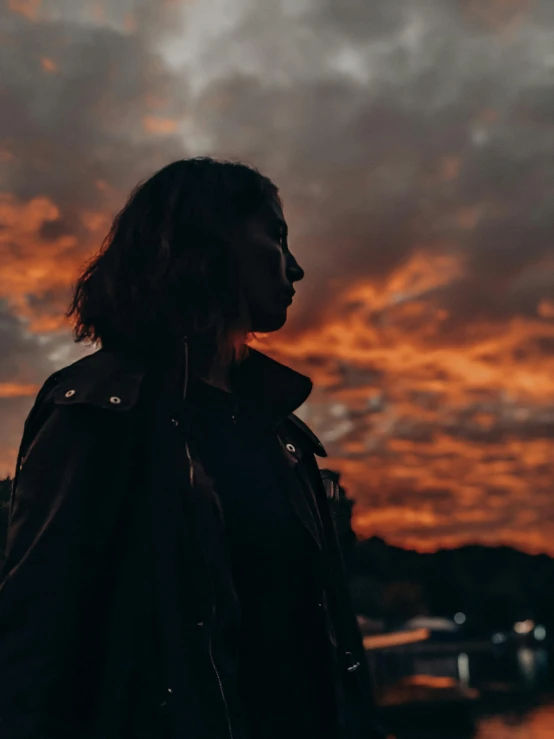 a woman standing in front of a sunset, by Emma Andijewska, black and orange coat, gloomy lights in the sky, profile image, red clouds