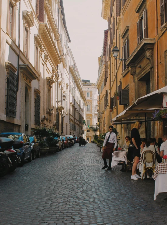 a narrow cobblestone street lined with parked cars, a photo, pexels contest winner, neoclassicism, people outside eating meals, profile image, young handsome pale roma, gif