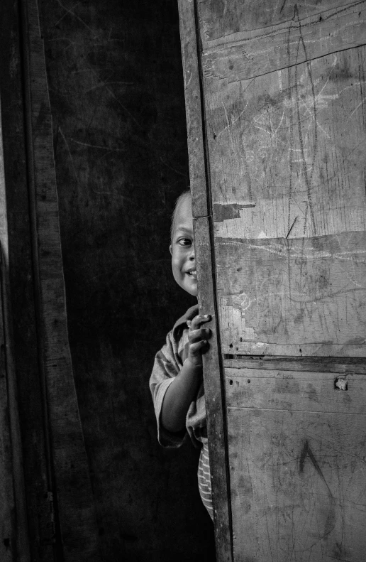a black and white photo of a child peeking out of a door, by Joze Ciuha, pexels contest winner, with a orientalist smileful face, stacked image, humanity, looking outside