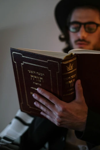a man sitting in a chair reading a book, an album cover, by Emanuel Witz, pexels, unilalianism, jewish young man with glasses, holding holy symbol, a person standing in front of a, islamic