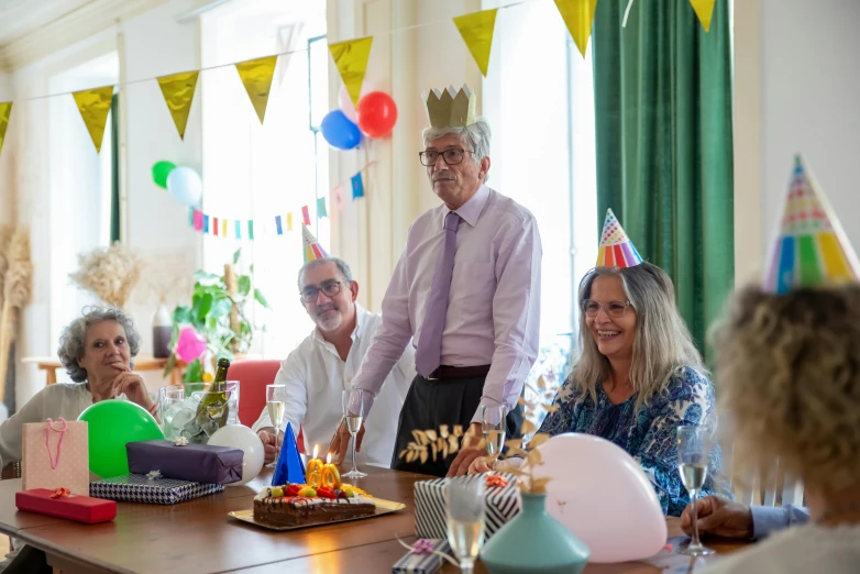 a group of people sitting around a table with a cake, an 80 year old man, party hats, background image, colour photograph