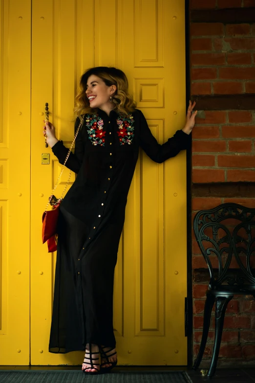 a woman standing in front of a yellow door, inspired by Eva Gonzalès, red gold and black outfit, embroidered shirt, wearing a long dress, press shot