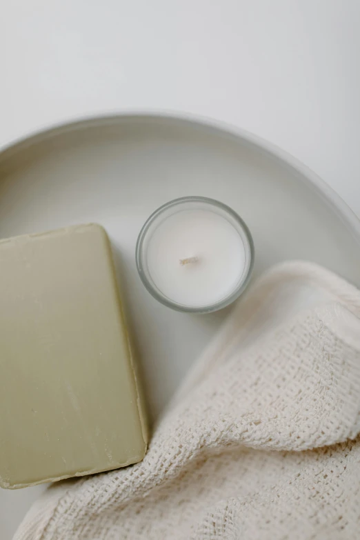 a soap bar sitting on top of a plate next to a candle, sustainable materials, cream white background, detailed product image, bathtub