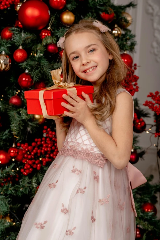 a little girl holding a present in front of a christmas tree, a picture, shutterstock, dressed in a pink dress, thumbnail, high resolution image, small