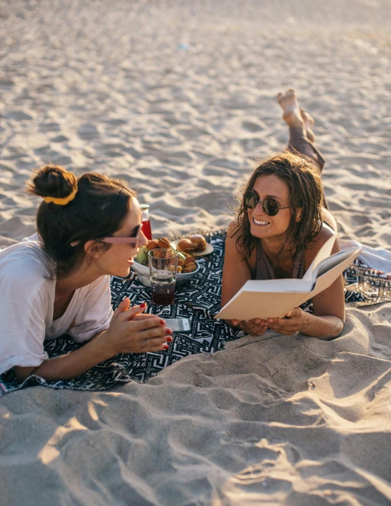 a couple of women laying on top of a sandy beach, pexels contest winner, holding a book, smiling at each other, gif, people outside eating meals