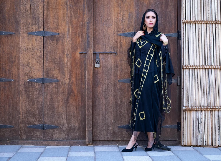 a woman standing in front of a wooden door, arabesque, wearing black clothes and cape, dubai, facebook post, portrait image