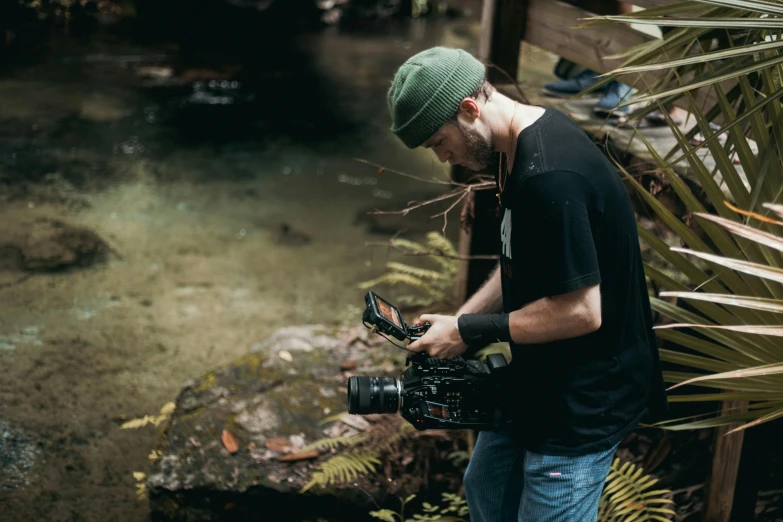 a man standing next to a river holding a camera, film production, in a jungle environment, worksafe. cinematic, instagram picture