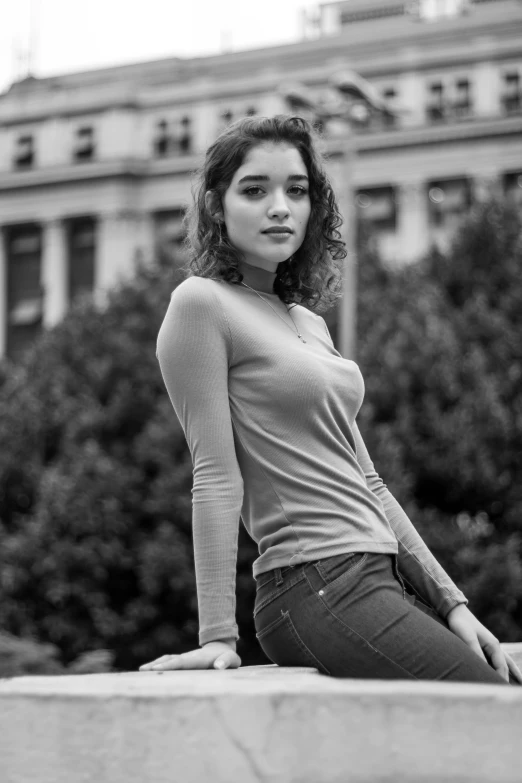 a woman sitting on a ledge in front of a building, a black and white photo, inspired by Ruth Orkin, renaissance, 🤤 girl portrait, tight shirt, portrait sophie mudd, curly