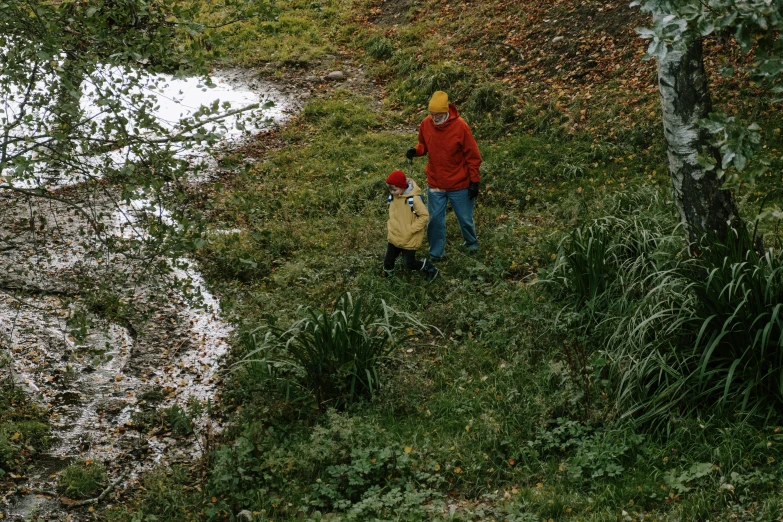 a couple of people that are standing in the grass, by Attila Meszlenyi, pexels, small river on the ground, yellow raincoat, with a kid, a high angle shot