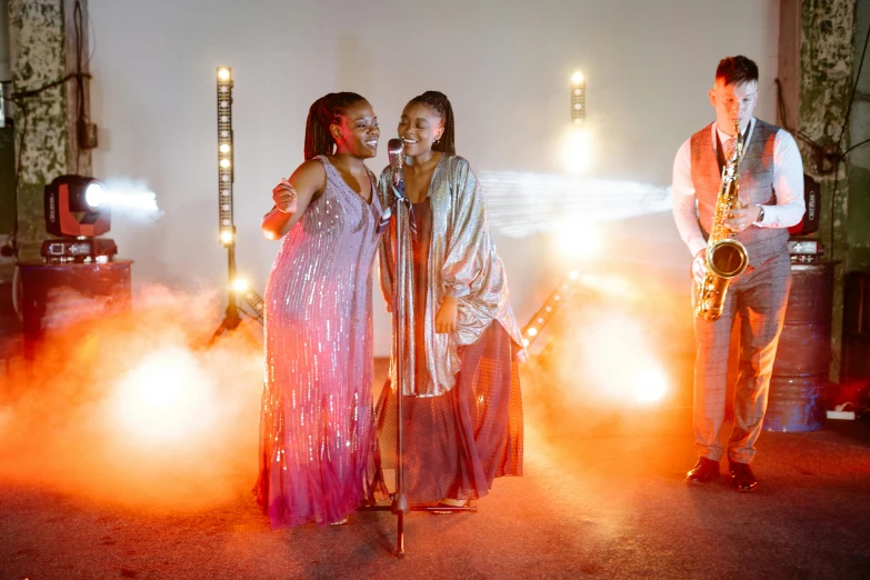 a group of people standing next to each other on a stage, an album cover, pexels, happening, two women kissing at a carnival, wearing long silver robes, studio lit, performing a music video