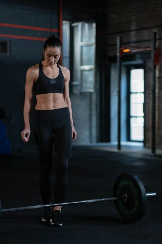 a woman standing next to a barbell in a gym, a portrait, by Paul Bird, pexels contest winner, wearing crop top, walking, concerned, manuka