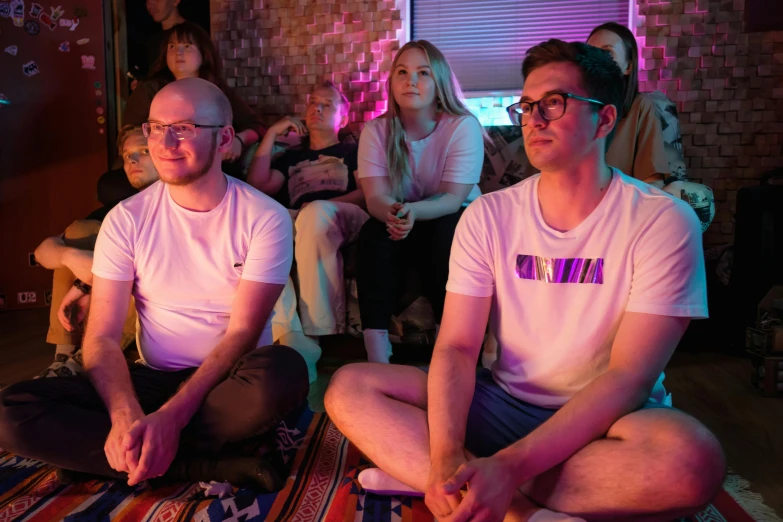 a couple of men sitting on top of a rug, tv screens in background, bisexual lighting, audience, meditating