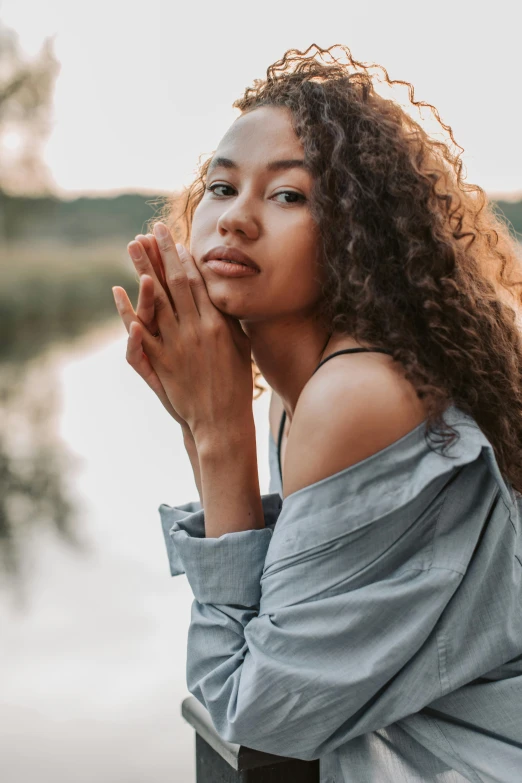 a woman sitting next to a body of water, trending on pexels, renaissance, with curls, mixed race woman, soft glow, crossed arms