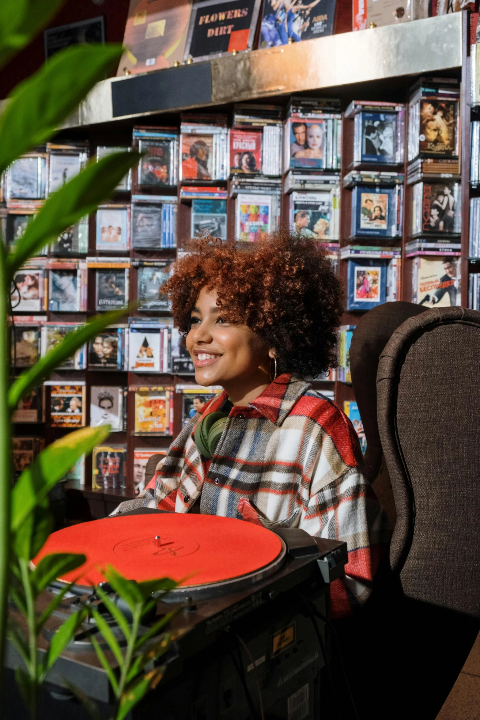 a woman sitting at a table in front of a wall of dvds, an album cover, trending on pexels, funk art, curly red hair, turntables, official store photo, portrait of a museum art curator