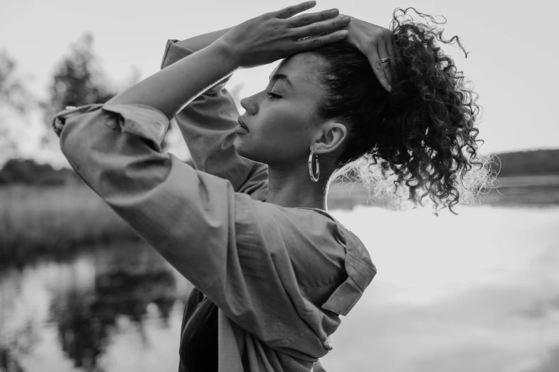 a woman standing next to a body of water, a black and white photo, by Emma Andijewska, pexels contest winner, curly bangs and ponytail, african american elegant girl, greeting hand on head, woman in streetwear