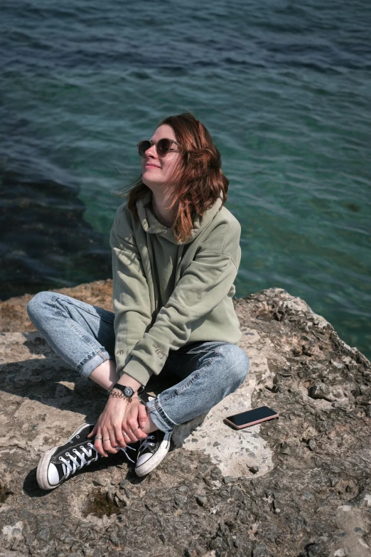 a woman sitting on top of a rock next to the ocean, inspired by Elsa Bleda, trending on pexels, happening, casual green clothing, with sunglass, wearing sweatshirt, pale skin