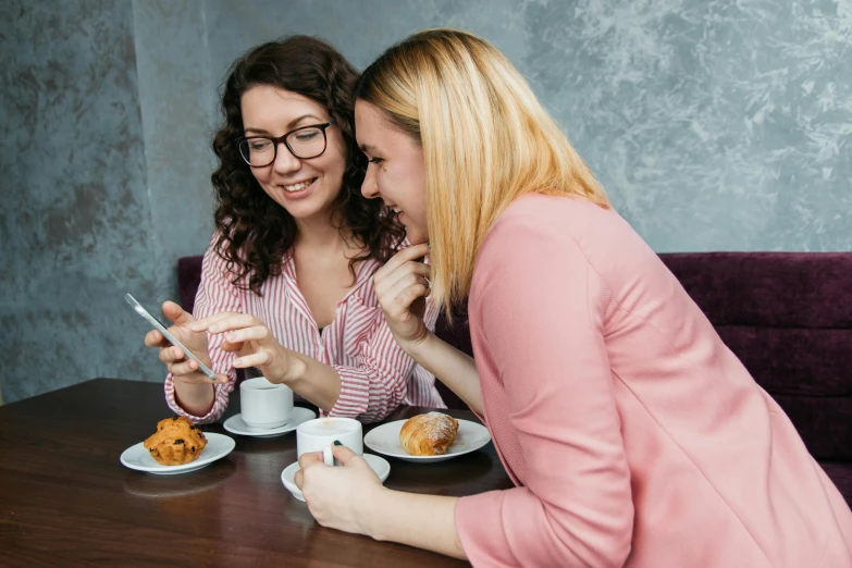 two women sitting at a table looking at a cell phone, trending on pexels, aussie baristas, avatar image, eating cakes, one blonde and one brunette