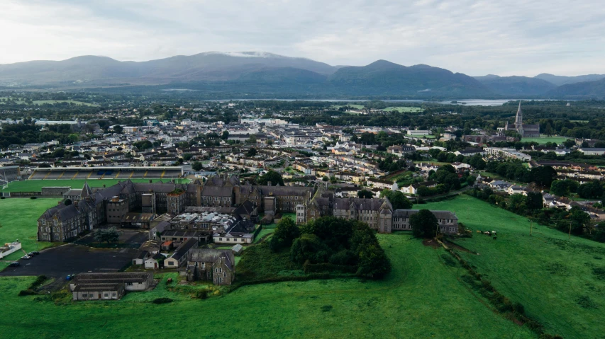 a large building sitting on top of a lush green field, pexels contest winner, irish mountains background, aerial view of a city, thumbnail, small town surrounding
